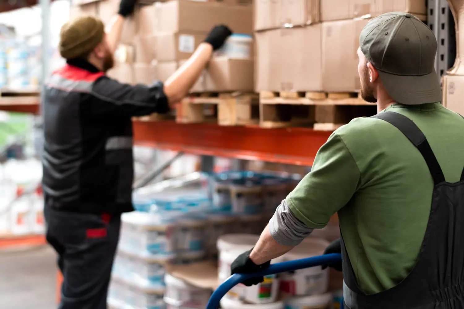 Two warehouse workers collaborate to stock shelves; one pulls a hand truck in the foreground while another retrieves a box from a high rack in the background.