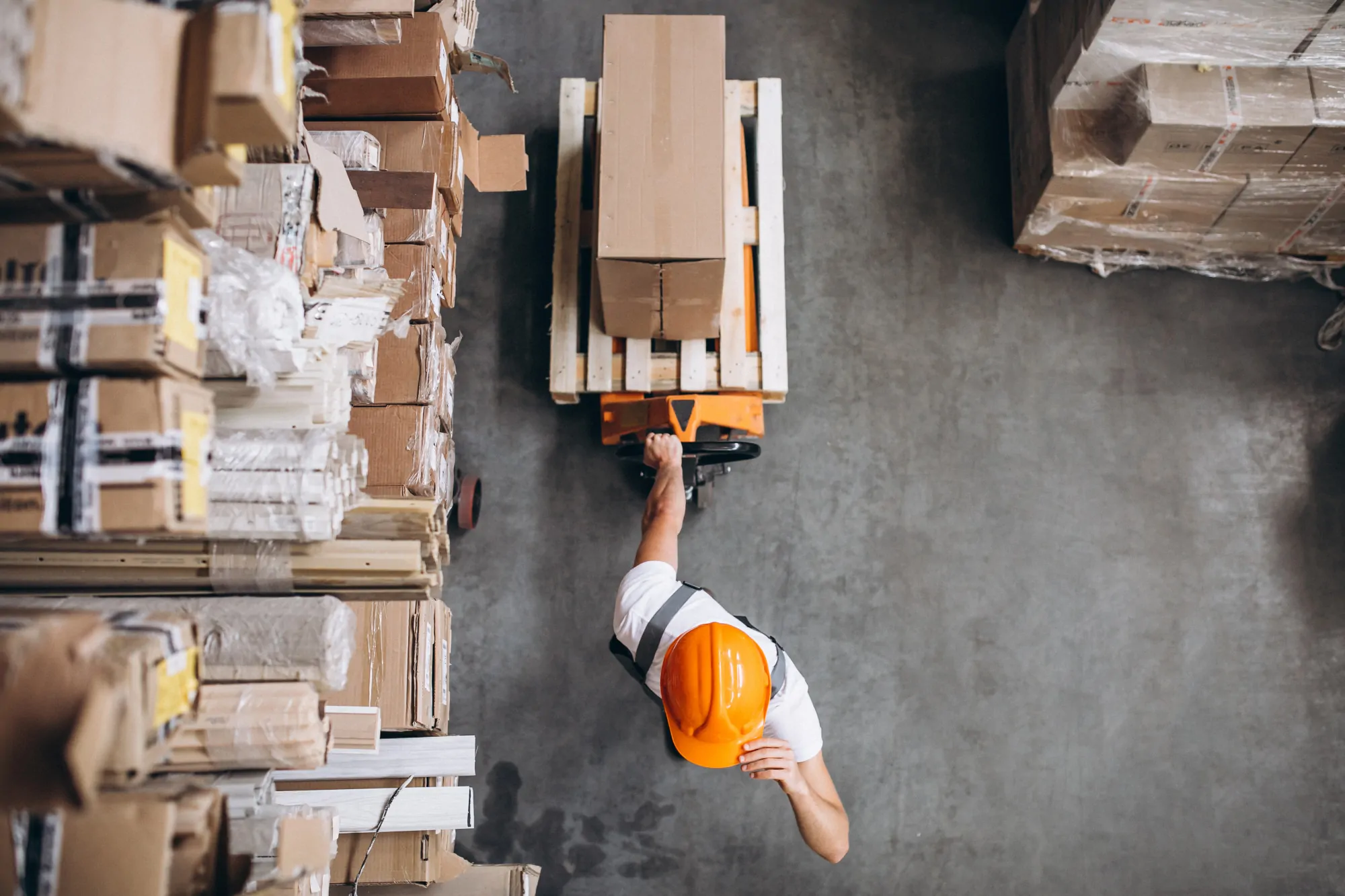 Top-down aerial view of a worker in an orange hard hat pulling a pallet jack with a box on it between tall warehouse shelving units.
