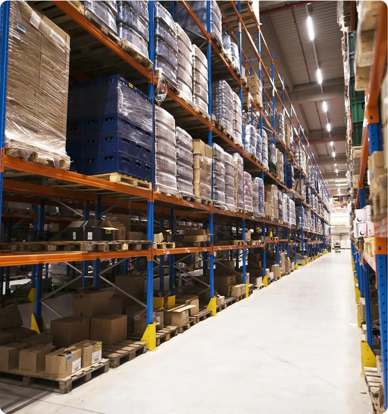 A long, clean aisle in a large warehouse, flanked by towering industrial shelves packed with pallets of inventory reaching toward the high ceiling.