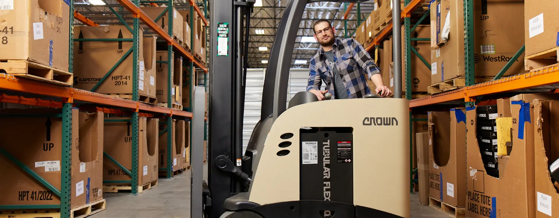 A man in a plaid shirt and glasses operates a Crown stand-up forklift in a warehouse aisle between tall racks stacked with large cardboard boxes.