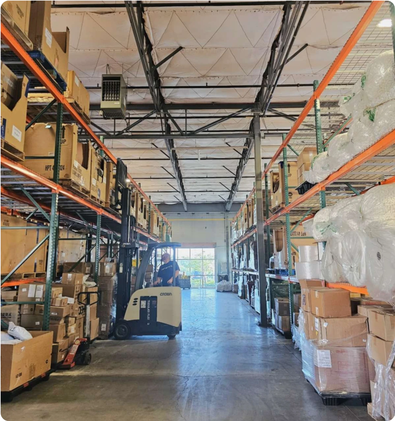 A view down a long warehouse aisle where a person is operating a forklift, with tall industrial shelves stacked high with boxes and inventory on both sides.