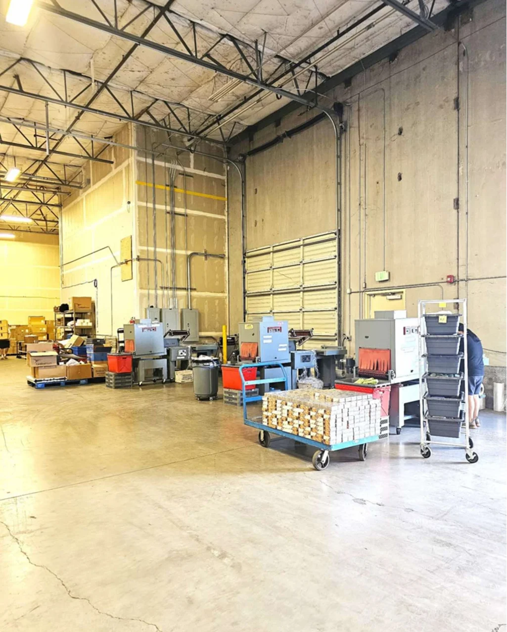 A wide view of a brightly lit warehouse section equipped with various industrial machines for preparing goods, including shrink-wrap equipment and carts of bundled products.
