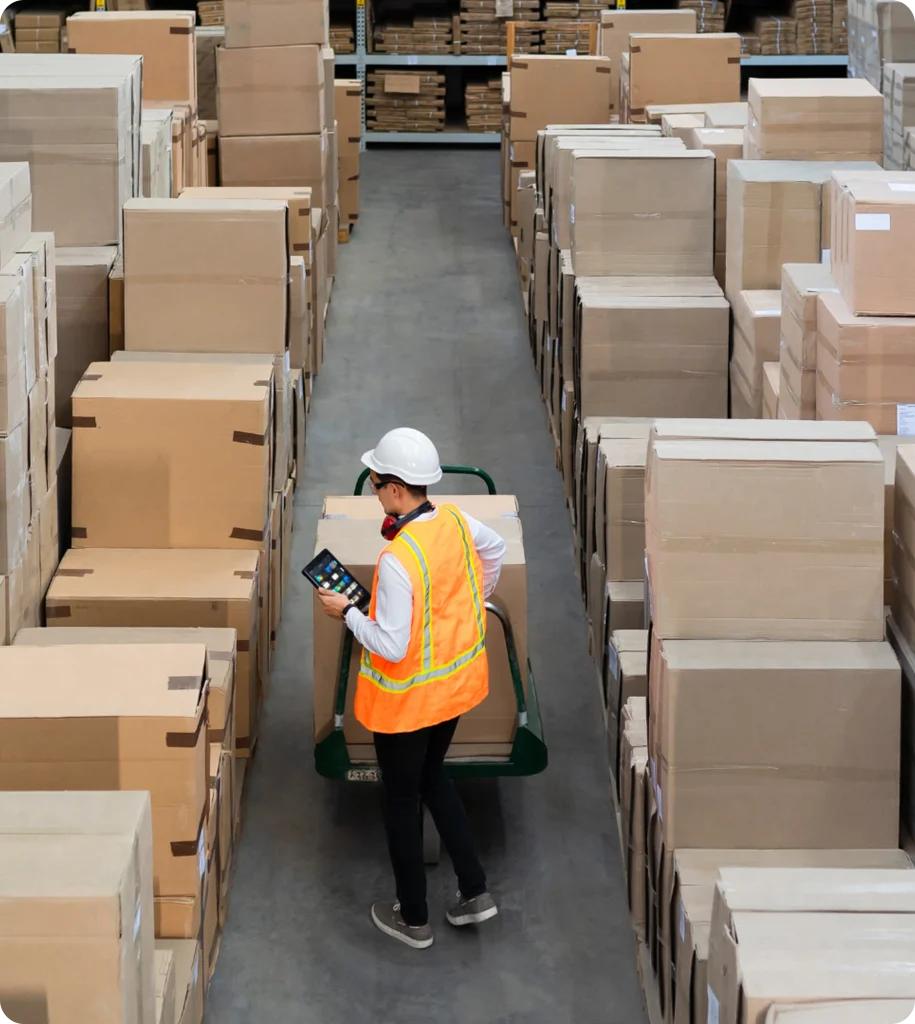 A high-angle view of a warehouse worker wearing a hard hat and orange safety vest, looking at a tablet while pushing a hand truck down a narrow aisle stacked high with cardboard boxes.